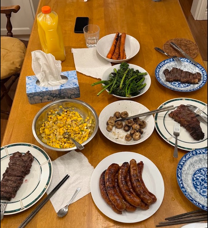 The full dinner spread: steaks, broccolini, carrots, mushrooms, corn, and sausages on the table