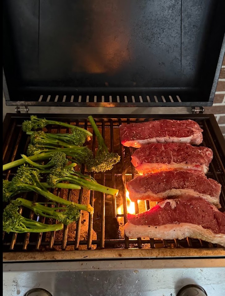 NY strip steaks and broccolini cooking over flames on the grill