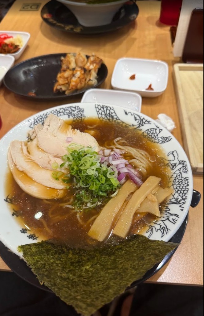 Bowl of cold Hakata ramen with sliced chicken, green onion, red onion, bamboo shoots, and nori, with chicken gyoza in the background