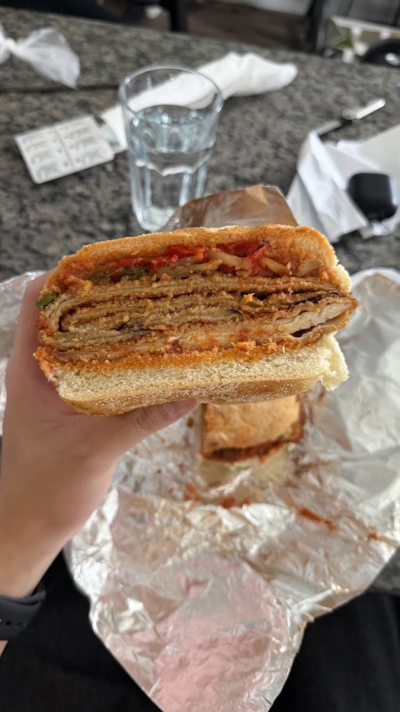 Cross-section of a chicken and eggplant sandwich from Uno Mustachio with crispy breaded layers, roasted peppers, and toppings on a bun